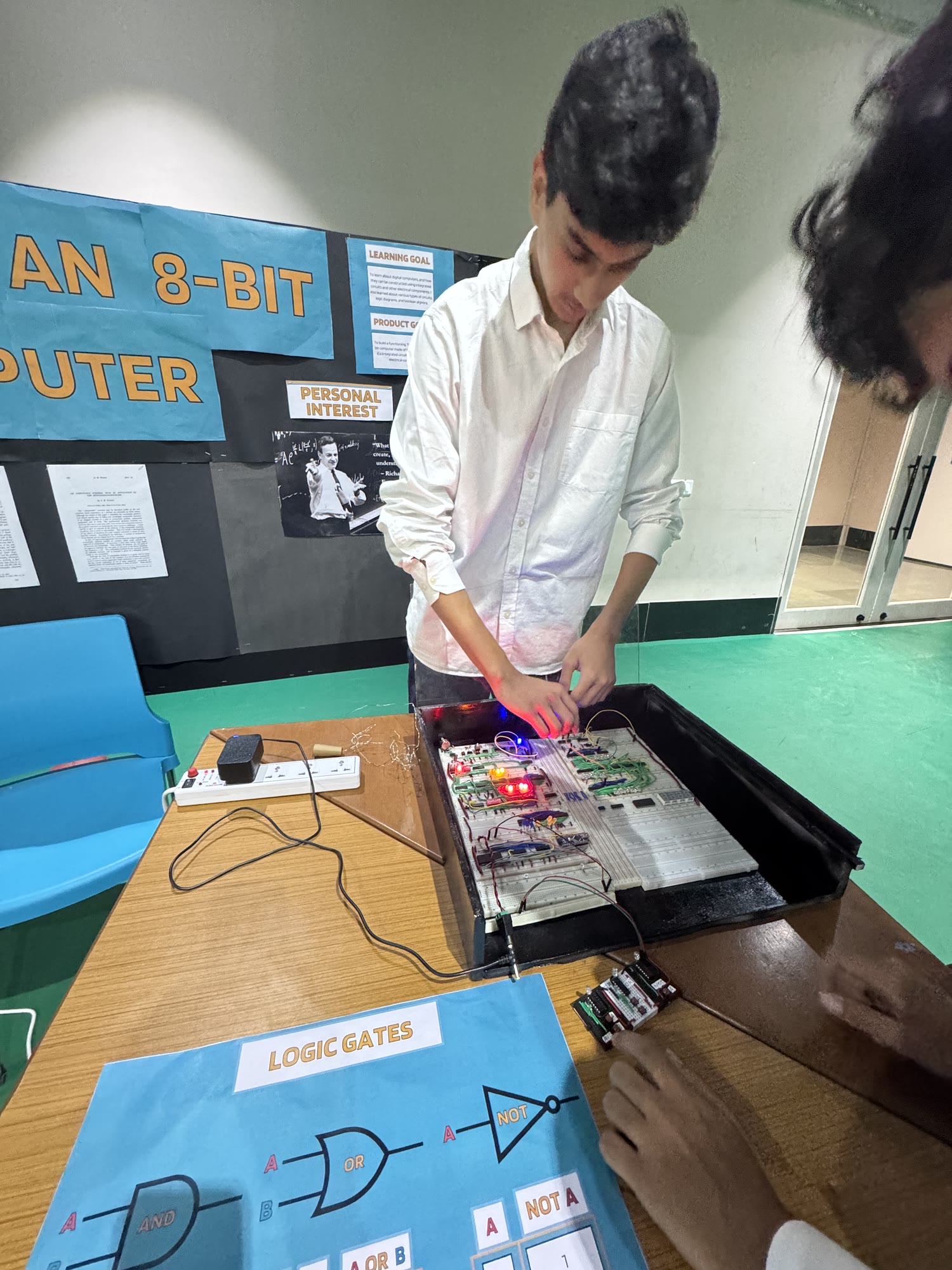 Arjun wires a breadboard circuit with lit LEDs. A poster behind him reads "An 8-bit computer", and a chart of logic gates lies on the table.