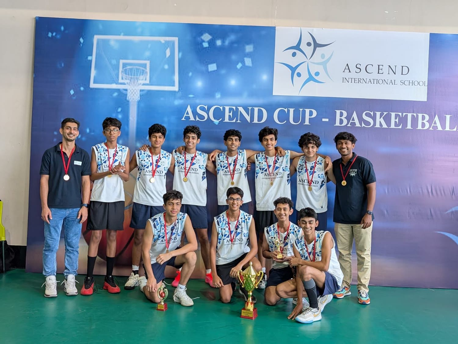 The Ascend basketball team and coaches, wearing medals, kneel and stand around a trophy in front of an Ascend Cup Basketball banner.