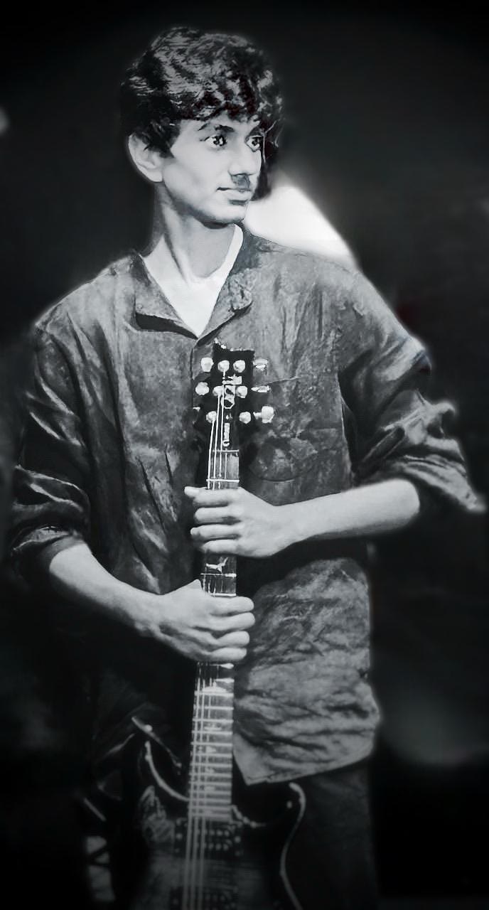 Black and white portrait of Arjun holding an electric guitar upright.
