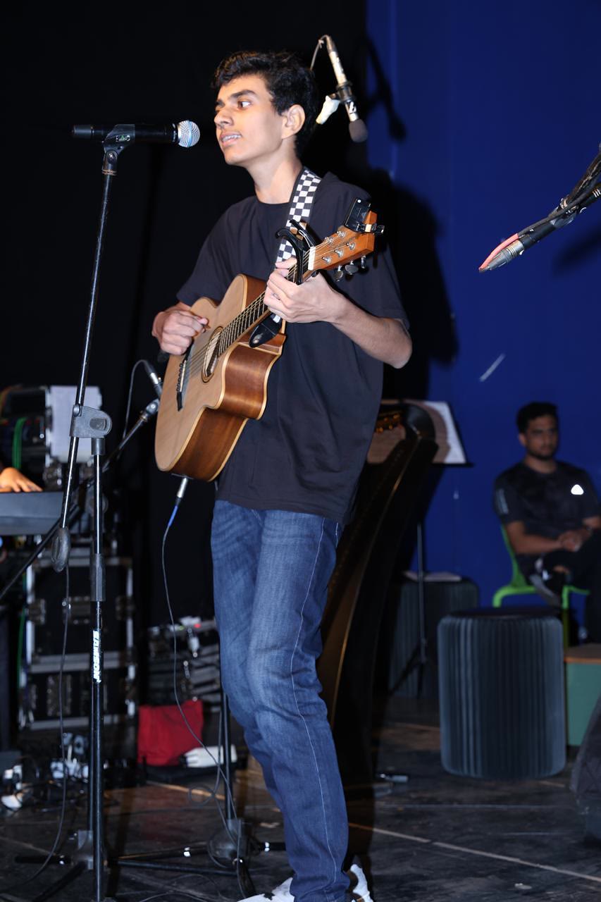 Arjun sings and plays an acoustic guitar at a microphone on a stage with a blue backdrop.