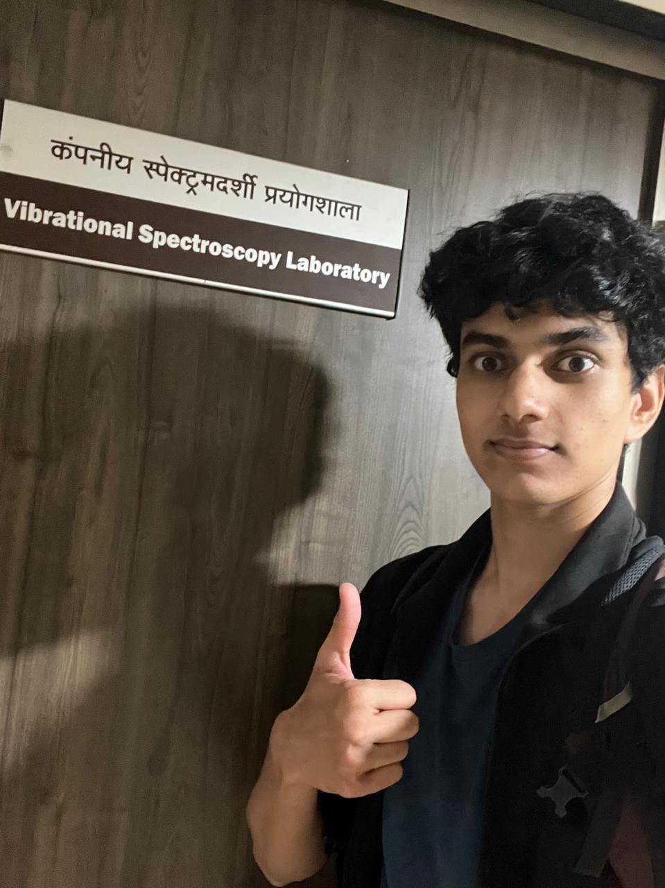 Arjun gives a thumbs up beside the door sign for the Vibrational Spectroscopy Laboratory.