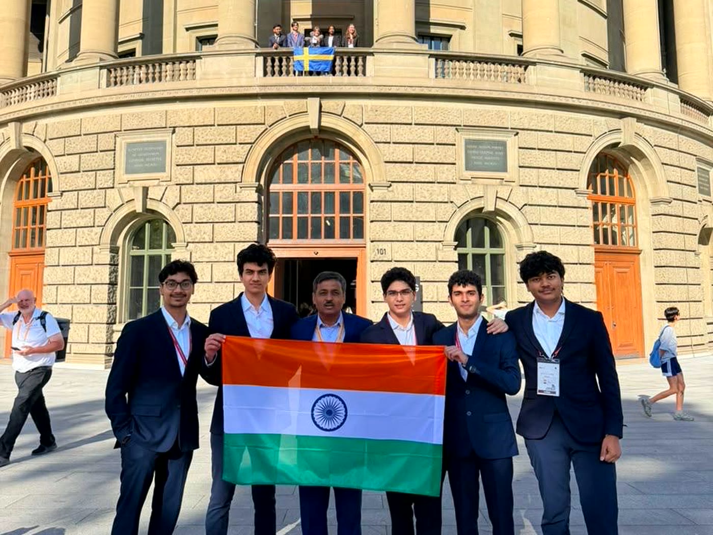 Five students and an adult in blazers hold the Indian flag outside a stone building with arched windows.