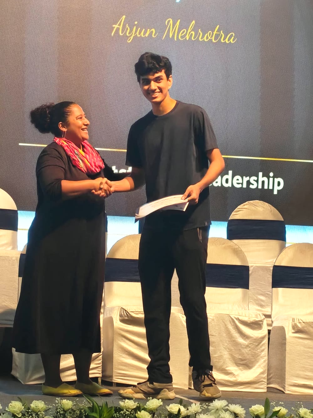 Arjun shakes hands on stage and holds a certificate. His name is on the screen behind him.
