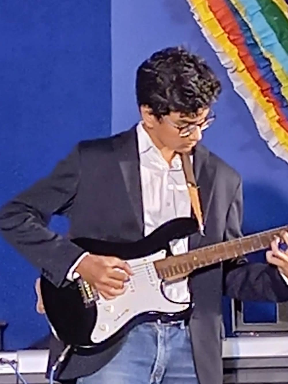 Arjun plays a black electric guitar on a stage with a blue backdrop.