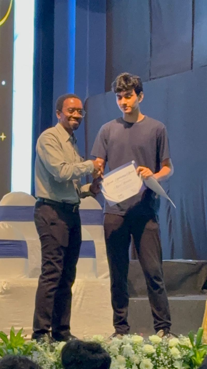 Arjun receives a certificate on a stage with flowers and a blue backdrop.