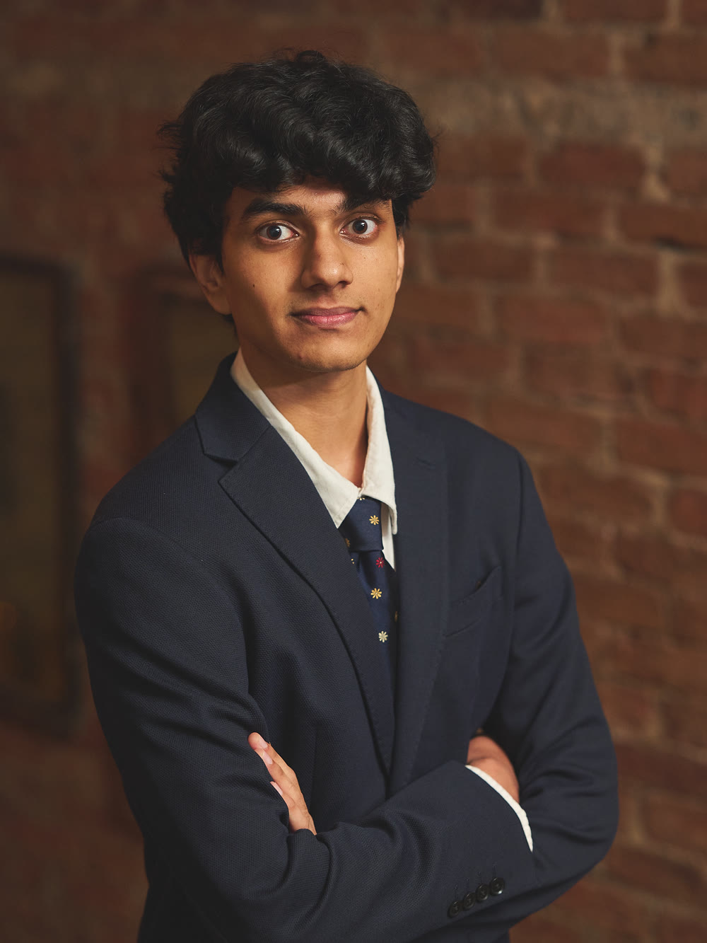 Arjun Mehrotra in a navy blazer and tie, arms folded, in front of a brick wall.